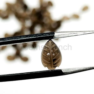 May include: A brown, leaf-shaped gemstone is held by tweezers against a white background. The gemstone has carved details resembling leaf veins. The tweezers are black and the gemstone is a light brown color.