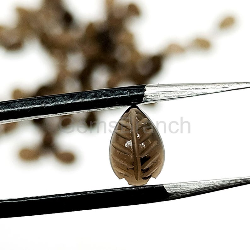 May include: A brown, leaf-shaped gemstone is held by tweezers against a white background. The gemstone has carved details resembling leaf veins. The tweezers are black and the gemstone is a light brown color.