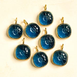 Könnte beinhalten: Acht blaue Edelsteinanhänger mit floralem Design. Jeder Anhänger hat einen goldfarbenen Rahmen und eine Schlaufe zum Aufhängen. Die Anhänger sind quadratisch mit abgerundeten Ecken.
