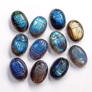 May include: A selection of oval labradorite scarab beetle carvings. The stones exhibit iridescent blue, green, and gold tones. Each beetle is intricately carved with detailed lines, creating a textured surface. The carvings are arranged on a white background.