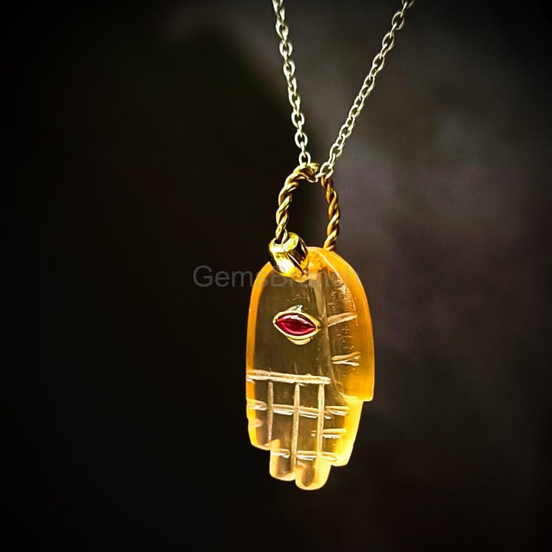 K&ouml;nnte beinhalten: Eine goldfarbene Halskette mit einem handf&ouml;rmigen Anh&auml;nger. Der Anh&auml;nger ist durchscheinend bernsteinfarben mit einem roten Akzent. Die Kette ist filigran und vor einem dunklen Hintergrund.