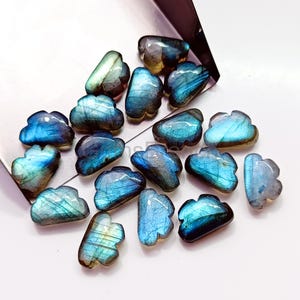 May include: A selection of cloud-shaped labradorite gemstones. The stones exhibit blues, greens, and greys, with iridescent flashes. The polished stones are arranged on a white surface, with a metallic container in the background.