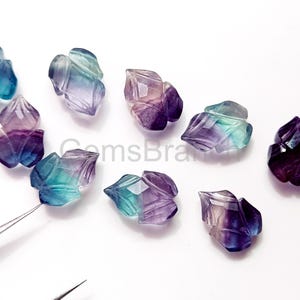 Puede incluir: Una colecci&oacute;n de hojas de piedras preciosas de fluorita talladas en tonos morado, azul y verde. Las piedras preciosas est&aacute;n facetadas y pulidas, con vetas visibles. Las hojas est&aacute;n dispuestas sobre una superficie blanca.