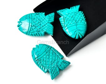 May include: Three turquoise fish-shaped carvings with intricate etched details. The carvings are a vibrant teal color with dark veining, and are displayed against a white background and a black box. The carvings are likely gemstones or decorative elements.