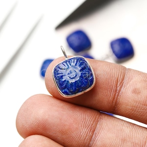 Può includere: Un ciondolo quadrato blu scuro con un motivo floreale intagliato sulla superficie. Il ciondolo è incastonato in una cornice di metallo color argento e ha un piccolo anello nella parte superiore per appenderlo. Sullo sfondo sono visibili altri ciondoli simili.