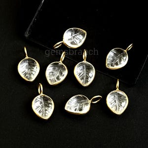 May include: Seven leaf-shaped pendants with a clear, faceted design and gold-colored metal trim. The pendants have a loop at the top for hanging. The background is black.