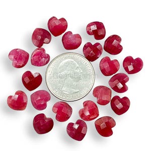 May include: A collection of faceted, heart-shaped gemstones in varying shades of red and pink. A US quarter coin is included for size reference. The gemstones are scattered on a white surface, showcasing their polished surfaces and unique shapes.