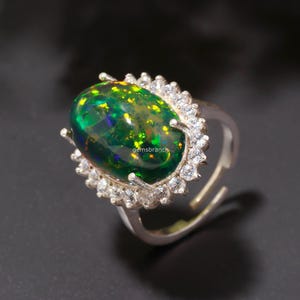 May include: A silver ring featuring a large, oval-cut black opal gemstone with vibrant green and blue flashes. The opal is surrounded by a halo of small, clear gemstones. The ring is adjustable and has the text "gemsbranch".