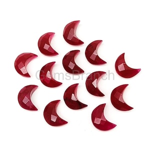 May include: A collection of faceted, crescent-shaped gemstones in a deep red hue. The gemstones are arranged on a white surface, showcasing their polished surfaces and geometric cuts. The overall theme is jewelry making.