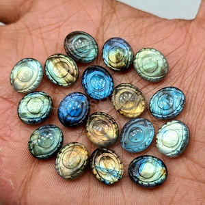 May include: A collection of oval-shaped labradorite gemstones in various colors, including blue, green, and gold. Each stone has a unique pattern and iridescent sheen, with carved details. The gemstones are displayed on a hand.