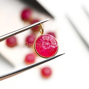 Puede incluir: Un colgante redondo de color rubí con un bisel dorado es sujetado por unas pinzas. El colgante tiene un diseño grabado en su superficie. Varias otras piedras de color rubí son visibles en el fondo, fuera de foco.