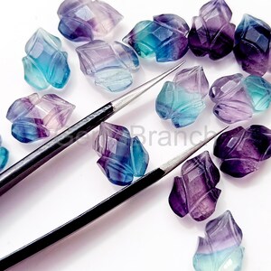 Puede incluir: Un conjunto de gemas de fluorita facetadas en forma de flor, en tonos azules, morados y turquesas. Las piedras preciosas est&aacute;n dispuestas sobre un fondo blanco. Tambi&eacute;n se ven dos herramientas plateadas, posiblemente para la fabricaci&oacute;n de joyas.