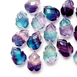 Puede incluir: Una colecci&oacute;n de gemas de fluorita talladas en forma de flores. Las gemas muestran una gama de colores, incluyendo p&uacute;rpura, azul y verde. Las superficies facetadas capturan la luz, creando un efecto brillante. Ideales para la creaci&oacute;n de joyas o coleccionar.