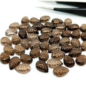 May include: A collection of brown, leaf-shaped gemstones. Each gem is carved with detailed leaf vein patterns. The gems are scattered on a white surface, with a pair of tweezers in the background.