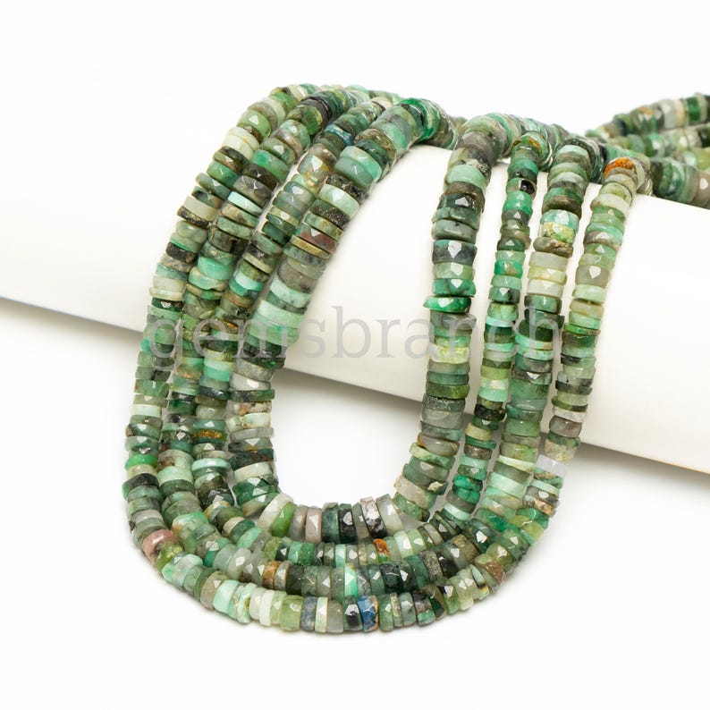 May include: A strand of green gemstone beads, with a faceted, irregular shape. The beads are strung on a white cord and are arranged in a loose, flowing pattern.
