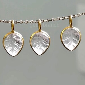 May include: Three leaf-shaped pendants with a clear, etched design, suspended from a silver chain. Each pendant has a gold-colored border and a loop for hanging. The pendants are displayed against a neutral background.