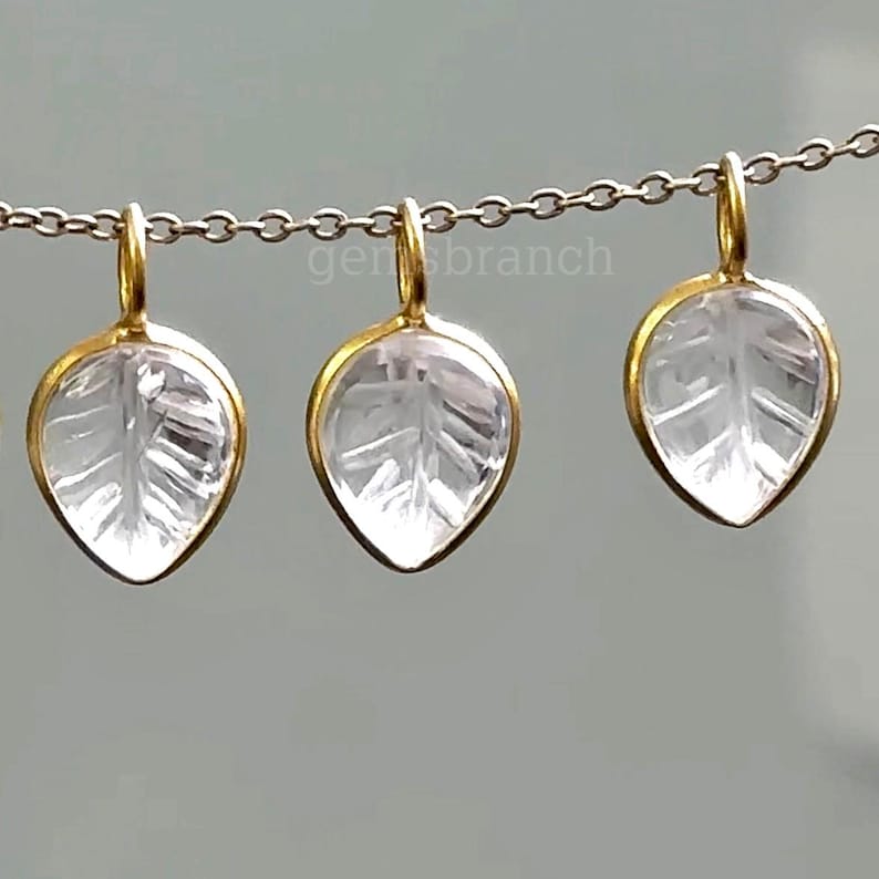 May include: Three leaf-shaped pendants with a clear, etched design, suspended from a silver chain. Each pendant has a gold-colored border and a loop for hanging. The pendants are displayed against a neutral background.
