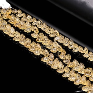 May include: Close-up of several strands of yellow gemstone beads. Each bead is carved in the shape of a leaf. The beads are strung together on a white string. The background is black.
