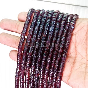May include: Multiple strands of dark red, faceted gemstone beads. The beads are small, square-cut, and strung together, creating a shimmering effect. The beads are held in a hand, showcasing their colour and texture. Suitable for jewellery making.