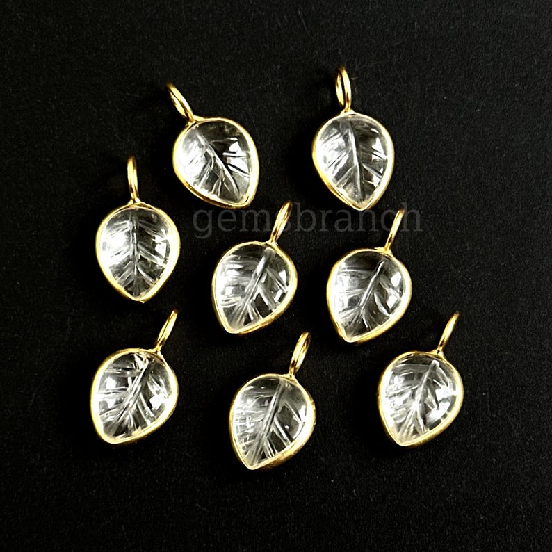 May include: Seven leaf-shaped pendants with clear, textured centers and gold-colored metal frames. Each pendant has a small loop at the top for hanging. The pendants are arranged on a black surface. The text "gem branch" is visible.