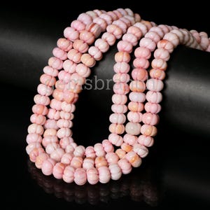 May include: A strand of pink shell beads, each bead is shaped like a small pumpkin. The beads are strung on a clear thread and are arranged in a circular pattern.