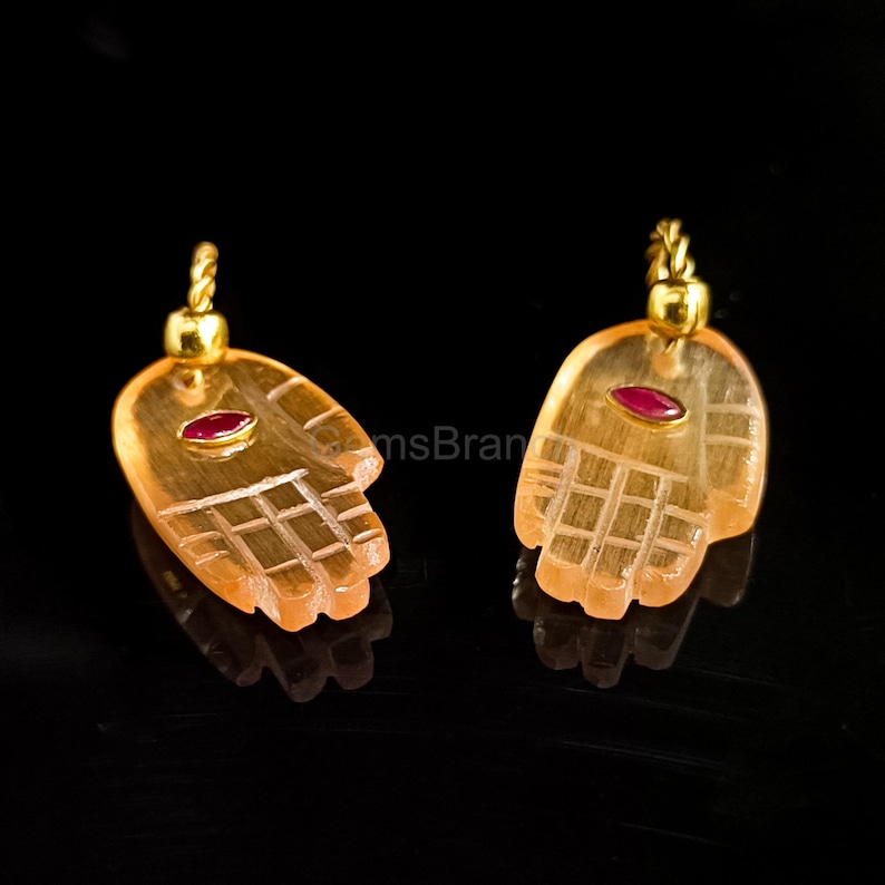 K&ouml;nnte beinhalten: Zwei handf&ouml;rmige Anh&auml;nger mit einem goldenen B&uuml;gel. Jeder Anh&auml;nger hat eine hellorange Farbe mit einem roten Edelstein in der Mitte. Das Handdesign hat ge&auml;tzte Linien, die die Finger und die Handfl&auml;che darstellen.