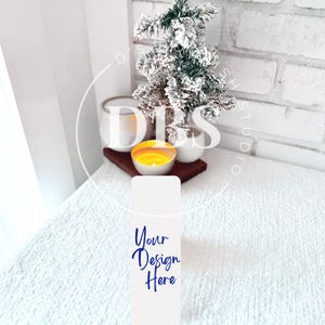 May include: A white rectangular product mockup with the text "Your Design Here" in blue script. A small, snow-covered Christmas tree and lit candle sit on a wooden tray, with a white brick wall in the background. The scene is set on a white, textured surface.
