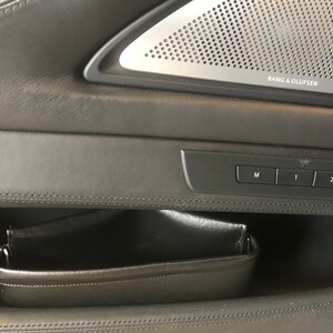 May include: A black leather car door panel with a storage compartment and a set of buttons labeled "M", "1", "2", and a seat icon. The panel also features a speaker grille with the text "Bang & Olufsen".