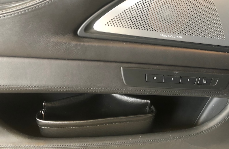 May include: A black leather car door panel with a storage compartment and a set of buttons labeled "M", "1", "2", and a seat icon. The panel also features a speaker grille with the text "Bang & Olufsen".
