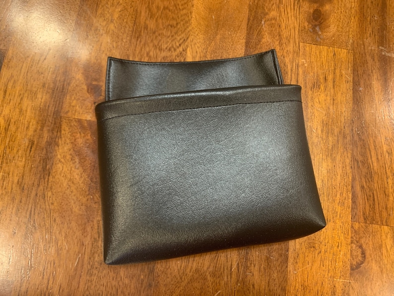 May include: A black leather pouch with a fold-over flap. The pouch is closed and sitting on a wooden surface.