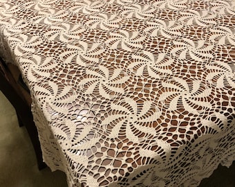 Könnte beinhalten: Eine cremeweiße, rechteckige Tischdecke mit einem komplizierten, sich wiederholenden Häkelmuster aus wirbelnden Windradmotiven. Das durchbrochene Design erzeugt eine zarte, spitzenartige Textur, die die gesamte Oberfläche bedeckt.