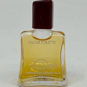 May include: A small, clear glass perfume bottle filled with amber liquid, featuring a dark maroon rectangular cap. The front of the bottle is printed with the text EAU DE TOILETTE, Osé, Parfums Guy Laroche Paris, and GUSTAV PARIS.