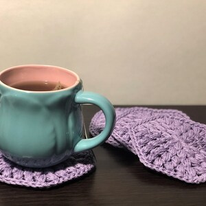 May include: A teal mug with a pink rim filled with tea sits on a purple crocheted coaster. Three additional purple crocheted coasters are in the background.