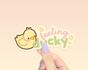 Puede incluir: Una pegatina de pato amarillo con el texto "feeling ducky" en letras rosa y verde. La pegatina tiene un contorno blanco y pequeños detalles de flores.