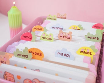 May include: A pink plastic storage box with dividers holds a collection of colorful index cards with cute animal and food illustrations. Each card has a unique number, such as "MS001" and "MV002".