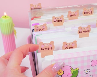 May include: A set of light brown bear-shaped paper index tabs attached to the top edges of white notebook pages. Each tab features a cartoon bear face with pink cheeks and handwritten text including BUMBIG, LVOOI, WCOOI, MM001, SPOOI, and SHOOS.