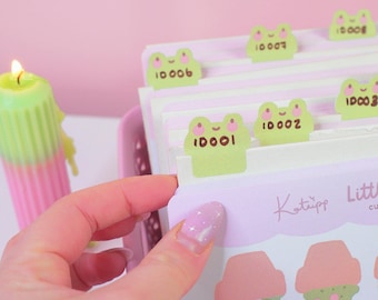 May include: A set of green frog-shaped index tabs with numbers on them. The tabs are labeled "10001", "10002", "10003", "10006", "10007", and "10008". The tabs are on a white and pink background with the text "Kotiypo Little cu"