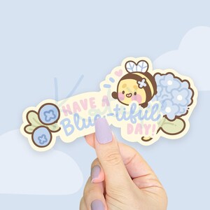 May include: A yellow and brown bee with a heart and a blue flower bouquet, with the text "Have a blue-tiful day!" and two blue blueberries on a white background.