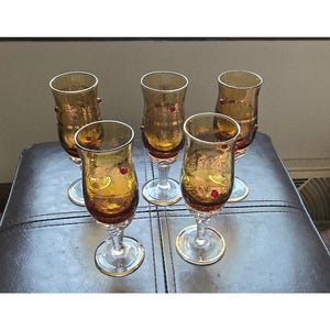 May include: Set of six amber glass stemmed drinking glasses with gold trim and red accents. The glasses are on a brown leather surface.