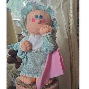 May include: A soft fabric doll with a peach-coloured face, blue eyes, and pink cheeks. The doll wears a light blue bonnet and dress featuring a small white floral pattern with white lace trim, a white bib, and a solid pink fabric piece draped over one side.