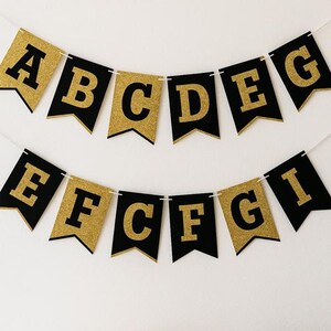 Puede incluir: Una pancarta de dos filas con banderines en forma de cola de golondrina con fondos negros y letras doradas con purpurina. La fila superior muestra las letras A, B, C, D, E, G y la inferior E, F, C, F, G, I, unidas por un cordón blanco.