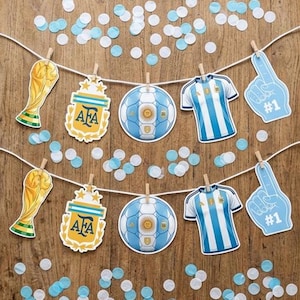 May include: A decorative banner featuring two rows of paper cutouts clipped to white strings. The cutouts include gold trophy replicas, AFA crests, soccer balls, blue and white striped jerseys, and blue foam fingers with the text #1.