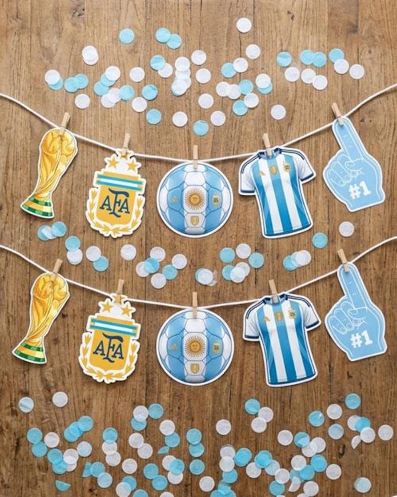 May include: A decorative banner featuring two rows of paper cutouts clipped to white strings. The cutouts include gold trophy replicas, AFA crests, soccer balls, blue and white striped jerseys, and blue foam fingers with the text #1.