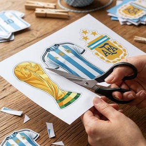 May include: A white sheet of paper featuring printed illustrations of a gold trophy, a light blue and white striped soccer jersey, and an AFA logo with three stars. A pair of black scissors is cutting out the trophy design from the paper sheet.