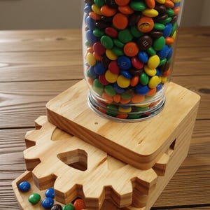 May include: A clear glass jar filled with colorful candies sits atop a wooden gear-shaped dispenser. The candies are various colors, including red, yellow, blue, and orange. The dispenser is made of light-colored wood.