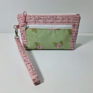 May include: A rectangular fabric wristlet pouch featuring a pink background with black cursive script text. The front includes a green pocket panel decorated with small pink rose illustrations and green leaves, secured by a white zipper and a wrist strap.