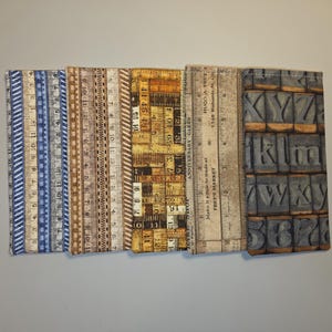May include: A collection of fabric-covered notebooks with ruler and letter block designs. The notebooks feature various colors, including blue, brown, and tan, with ruler markings and text. The last notebook has a design of letter blocks.