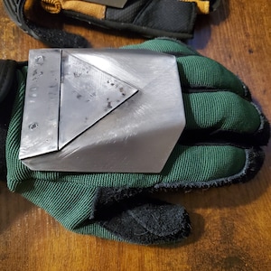 May include: A green work glove with a metal plate on the back of the hand. The plate has a triangular shape with a riveted edge.