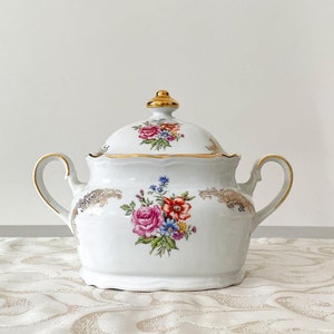 Może przedstawiać: Biała cukiernica z porcelany z pokrywką i dwoma uchwytami. Cukiernica jest ozdobiona kwiatowym wzorem w odcieniach różowego, czerwonego, żółtego i niebieskiego. Pokrywka i uchwyty są ozdobione złotem.