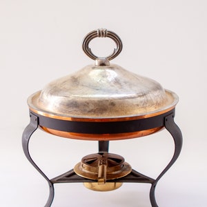 Puede incluir: Un plato calentador de plata con base de cobre y un soporte de metal negro. El plato calentador tiene una tapa redonda y abovedada con un asa decorativa.